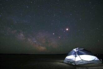 Starry night sky over illuminated tent in wilderness.
